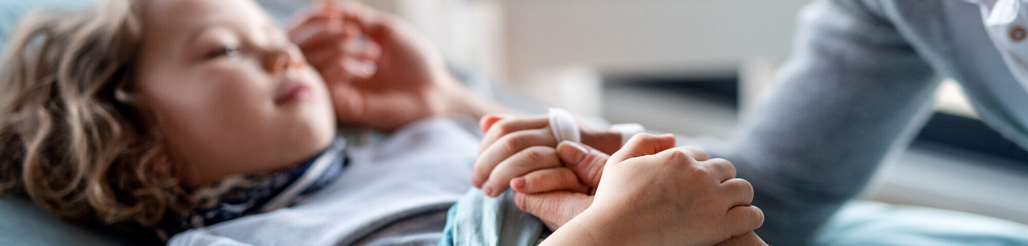 Kind liegt im Krankenbett und hält die Hand der Mutter