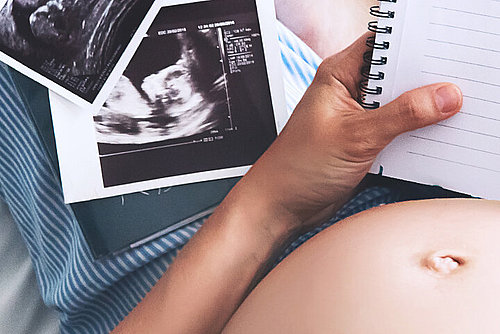 Hochschwangere Frau sitzt im Schneidersitz neben Ultraschalllbilder und schreibt in Notizbuch