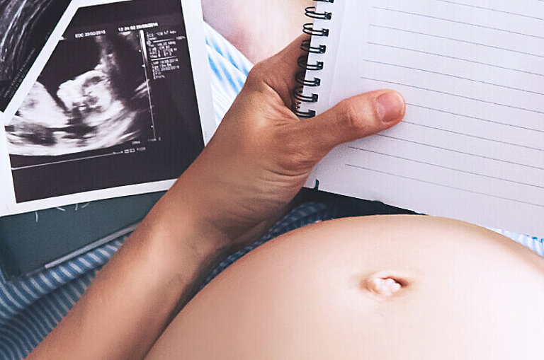 Hochschwangere Frau sitzt im Schneidersitz neben Ultraschalllbilder und schreibt in Notizbuch
